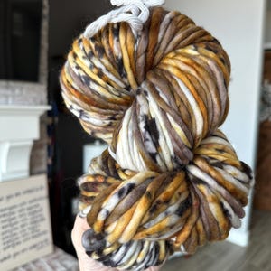 May include: A large, hand-dyed skein of super chunky yarn in a mix of colors. The yarn features shades of brown, gold, white, and black. The thick yarn is tied with a white string. This yarn is perfect for knitting or crocheting projects.