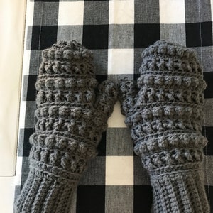 May include: A pair of gray crocheted mittens with a textured bobble stitch pattern. The mittens are on a black and white checkered surface.