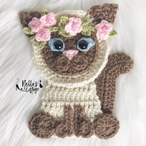 May include: A crocheted cat wearing a flower crown. The cat is brown and white and has blue eyes. The cat is sitting on a white fluffy surface.