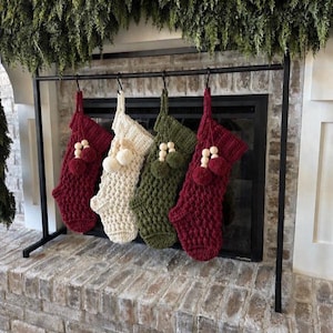 Peut inclure: Quatre chaussettes de Noël crochetées suspendues à une tringle en métal noir. Les chaussettes sont de couleur rouge foncé, crème et vert olive, avec un motif en relief et des perles en bois décoratives accompagnées de pompons en laine.