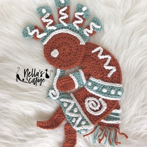 Pode incluir: Boneca Kokopelli de crochê feita com fio marrom, branco e turquesa. A boneca está de pé com os braços levantados e tem uma coroa com um design de espiral.