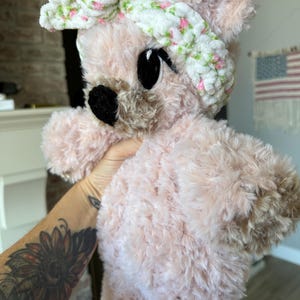 May include: A plush pink teddy bear with a white headband decorated with small pink and green floral accents. The bear has large black eyes and a black nose. The fur is soft and fluffy.
