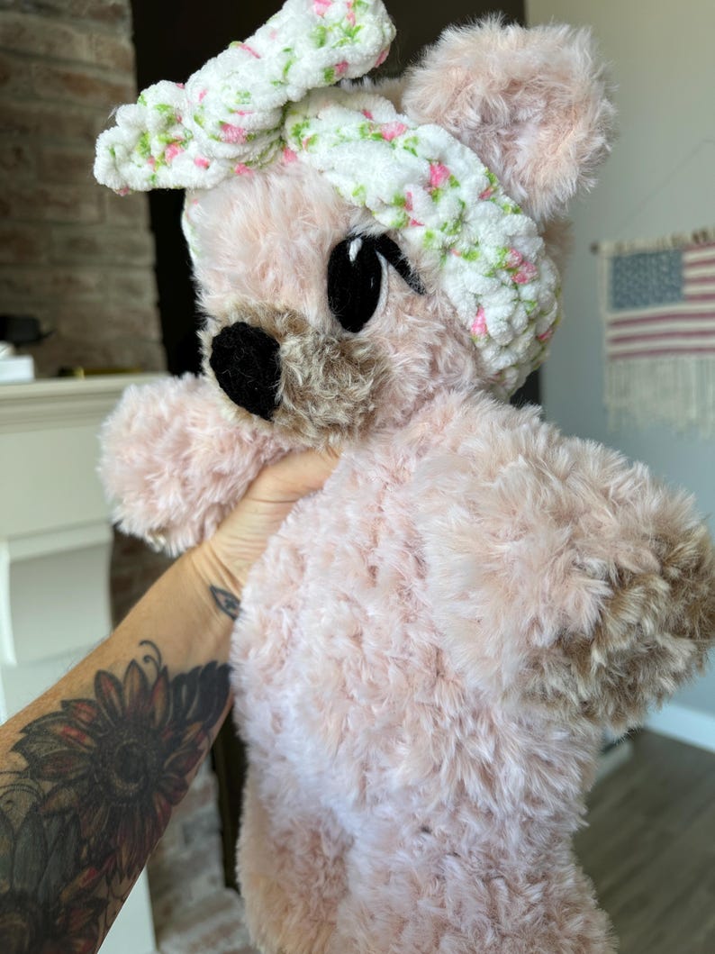 May include: A plush pink teddy bear with a white headband decorated with small pink and green floral accents. The bear has large black eyes and a black nose. The fur is soft and fluffy.
