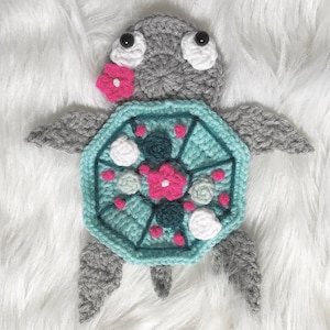 May include: A crocheted turtle with a teal shell decorated with white and pink flowers. The turtle has gray legs and a gray head with black eyes.