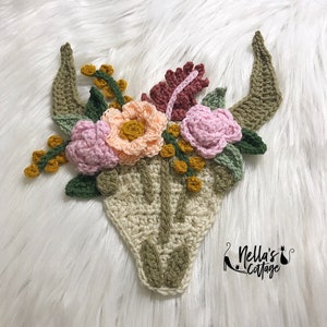 Puede incluir: Cráneo de toro tejido a crochet con adornos florales. El cráneo es beige con detalles verdes. Las flores son rosas, melocotón y amarillas con hojas verdes.