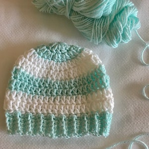 May include: A crocheted baby hat with white and light blue stripes. The hat is made with a soft yarn and has a simple design.