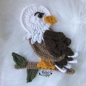 Könnte beinhalten: Ein gehäkelter Weißkopfseeadler mit braunen und weißen Federn, der auf einem Ast mit grünen Blättern sitzt. Der Adler hat einen gelben Schnabel und schwarze Augen. Der Ast hat eine weiße gehäkelte Basis und der Text "Nella's Cottage" ist sichtbar.