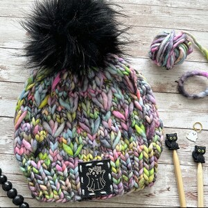 Op de afbeelding: Een kleurrijke gebreide beanie met een zwarte pompon en een zwarte leren patch met een spookontwerp. De beanie is gemaakt met verschillende kleuren, waaronder roze, paars, blauw, groen en geel.