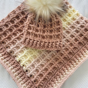 Może przedstawiać: Zestaw czapki i kocyka dla niemowlęcia wykonany na szydełku w odcieniach brązu i kremowego. Czapka ma puszysty biały pompon na górze.