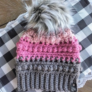 May include: A pink and gray crocheted beanie with a fluffy white pom pom. The beanie has a textured design with a gray band at the bottom. The beanie has a small black tag with the text "Bella's Cottage" on it.