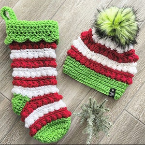 Peut inclure: Une chaussette de Noël en crochet rayée rouge, blanche et verte avec un revers vert et un pompon blanc sur le dessus. La chaussette est décorée de boules blanches et rouges. Un bonnet assorti en crochet rayé rouge, blanc et vert avec un revers vert et un pompon blanc sur le dessus est également présenté.