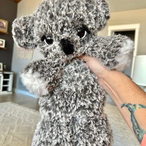 May include: A plush koala bear toy with gray and white fur. The koala has large, dark eyes, a black nose, and small, rounded ears. The toy is held up, showcasing its soft, fluffy texture.