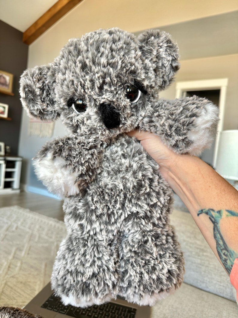 May include: A plush koala bear toy with gray and white fur. The koala has large, dark eyes, a black nose, and small, rounded ears. The toy is held up, showcasing its soft, fluffy texture.