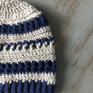 May include: A crocheted beanie hat with alternating stripes of beige and navy blue yarn.