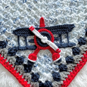 May include: A handmade crochet blanket featuring a blue, white, and gray pattern with a red and white airplane applique. The blanket has a red border and is made of soft yarn. The airplane has a propeller and is centered on the blanket.
