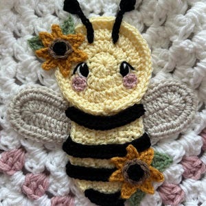 May include: A crocheted bee applique featuring a yellow head and body with black stripes, two beige wings, and two sunflowers with green leaves. The bee has black antennae, black eyes, and pink cheeks, centered on a white and pink crocheted background.