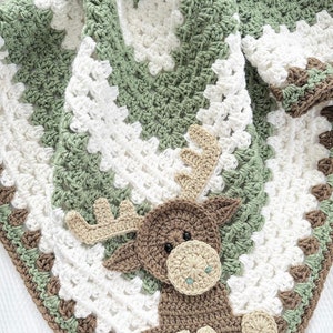 Puede incluir: Una manta de ganchillo hecha a mano con un diseño de alce. La manta es principalmente blanca con detalles en verde y marrón, y una aplicación detallada de alce en tonos marrones y beige. La manta es cuadrada y parece suave y acogedora.