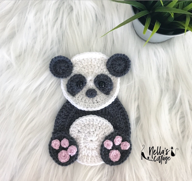 Peut inclure: Une applique en crochet de panda avec une fourrure noire et blanche, des coussinets de pattes roses et des yeux noirs. Le panda est assis sur une surface blanche et moelleuse.