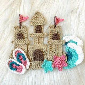 May include: A crocheted sandcastle with pink flags, brown windows, and a brown door. There are two pink flip flops, two turquoise starfish, and three turquoise and white waves.