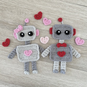 May include: Two crocheted robots, one gray and one pink, with hearts on their chests. The robots are holding hands and surrounded by pink and red hearts.