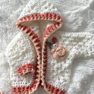 Puede incluir: Chaqueta de bebé de ganchillo blanco con una aplicación de flor rosa y ribete rosa. La chaqueta tiene capucha y está hecha con un punto delicado.