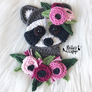 Könnte beinhalten: Ein gehäkelter Waschbär mit einem grau-weißen Gesicht und schwarzen Augen, der eine Krone aus rosa und weißen Blumen mit grünen Blättern trägt. Der Waschbär hält einen Strauß aus rosa und burgunderfarbenen Blumen mit grünen Blättern. Der Text "Nella's Cottage" ist in der rechten unteren Ecke des Bildes sichtbar.
