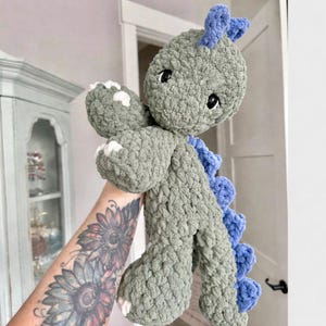May include: A handmade crochet dinosaur plushie in shades of gray and blue. The dinosaur has a textured, soft appearance with white accents on its paws and a row of blue spines down its back. The toy has black eyes and is designed to be cuddly.