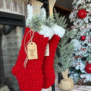 May include: Two red crocheted Christmas stockings with white trim and wooden name tags hang from a black metal rod. The stockings are decorated with faux greenery. A decorated Christmas tree is in the background.