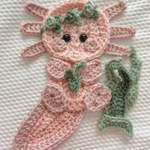 May include: A pink crocheted axolotl with green crocheted leaves and flowers. The axolotl has black eyes and a pink tail.