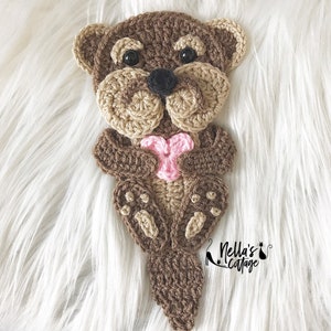 May include: A crocheted brown otter holding a pink heart. The otter is sitting on a white fluffy surface. The text "Nella's Cottage" is visible in the bottom right corner of the image.