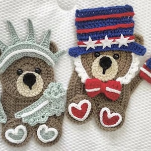 May include: Two crochet teddy bears, one dressed as the Statue of Liberty with a torch, the other as Uncle Sam with a flag. The bears are brown with white, red, blue, and yellow accents. The background is a white, textured surface.