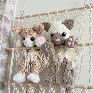 May include: Two handmade crochet cat dolls in a macrame hanging shelf. One cat is cream and brown, the other is orange and white. Both have large, expressive eyes and pink paw pads. The shelf is made of natural wood and cream-colored macrame.