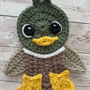May include: A handmade crochet duck with a green head, brown body, and yellow feet. The duck has black eyes with white highlights and a yellow beak. The wings are tan, and a white collar accents the neck. The duck is made of yarn.