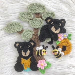 Puede incluir: Osos negros de crochet con ojos y narices marrones, sentados en un árbol de crochet marrón con hojas verdes. Hay dos abejas de crochet y una colmena de crochet con flores.