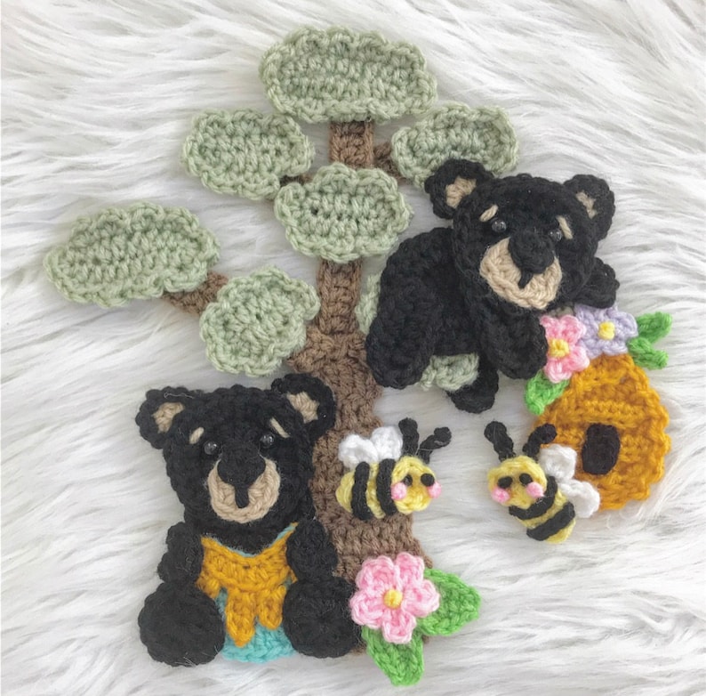 May include: Crocheted black bears with brown eyes and noses, sitting on a brown crocheted tree with green leaves. There are two crocheted bees and a crocheted beehive with flowers.