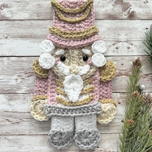 May include: A crocheted nutcracker ornament in pink, white, and gold. The nutcracker has a large hat, a white beard, and a detailed uniform.