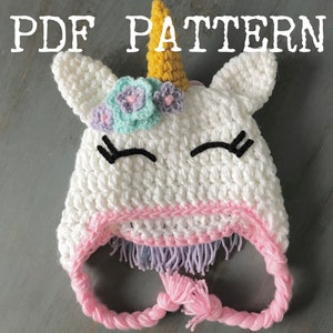 May include: White crochet unicorn hat with pink and purple trim, a yellow horn, and embroidered eyes. The hat has a long pink tie.