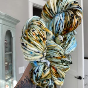 May include: A large skein of super chunky yarn in a mix of colors. The yarn features a blend of light blue, cream, yellow, orange, and dark green hues. The thick yarn is tightly wound and ready for crafting projects.