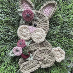 May include: A crocheted bunny with a floral crown and a bouquet of flowers. The bunny is made of tan yarn with pink accents on the ears and nose. The flowers are shades of pink and burgundy. The bunny is set against a backdrop of green foliage.