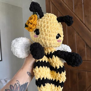 May include: A crocheted bee plush toy featuring a yellow and black striped body, black antennae, and white wings. The bee has large black safety eyes, pink stitched cheeks, and a yellow crocheted flower accessory attached to the side of its head.