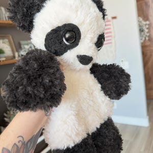 May include: A plush panda bear with black and white fur. The panda has black fur around its eyes, ears, arms, and legs. The bear's body is white, and it has large, black eyes and a small black nose. The panda is a stuffed animal.