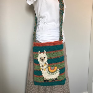 May include: A crocheted shoulder bag with a colorful striped design and a white llama applique. The bag has a long strap and is worn over the shoulder.