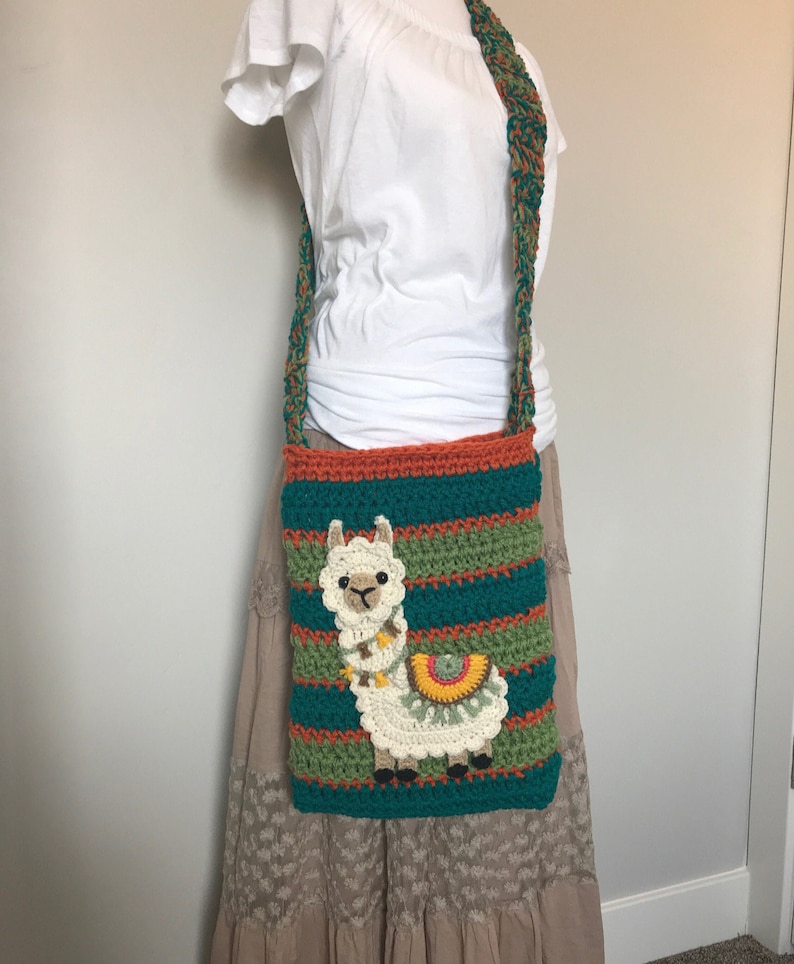 May include: A crocheted shoulder bag with a colorful striped design and a white llama applique. The bag has a long strap and is worn over the shoulder.