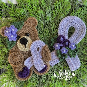 May include: A crocheted brown bear holding a purple ribbon with a flower embellishment. The bear is wearing a flower crown with purple and white flowers. The background is a green leafy plant.