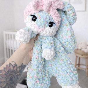 Puede incluir: Una muñeca de conejito de peluche hecha a mano en hilo azul claro, blanco y rosa suave. El conejito tiene orejas grandes y caídas, una cofia rosa y ojos negros de botón. La muñeca mide aproximadamente 30 cm de alto.