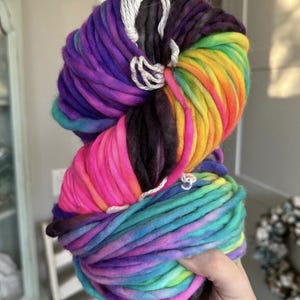 May include: A large, colorful skein of yarn in a rainbow of hues, including pink, purple, yellow, and teal. The thick yarn is held together with white string. This yarn is perfect for knitting or crocheting projects.