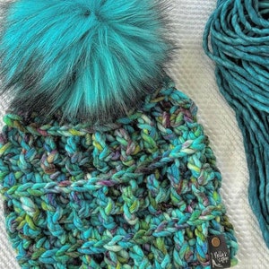 Peut inclure: Un bonnet en tricot bleu turquoise avec un pompon bleu turquoise duveteux. Le bonnet est fait avec un fil épais et présente un motif multicolore. Le bonnet est doté d'une étiquette en cuir marron avec le texte "Nella's Cottage" dessus.