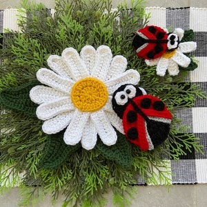 May include: A white crocheted daisy with a yellow center, surrounded by green foliage. Two red and black crocheted ladybugs are on the daisy and foliage.