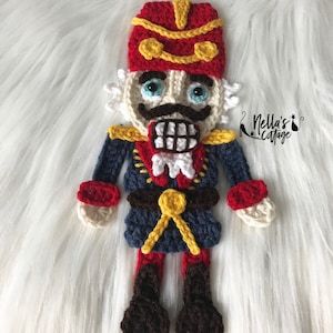 May include: A handmade crochet nutcracker doll in red, blue, and gold. The doll has a red hat with gold trim, a white face with blue eyes, and a black mustache. The nutcracker is wearing a blue jacket with gold trim and a red collar. The doll is approximately 8 inches tall.