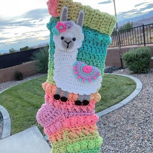 May include: A colorful crochet stocking with a white llama applique. The llama has a pink flower on its head and a pink and purple circle on its side. The stocking is made with a variety of colors, including pink, green, blue, and yellow.